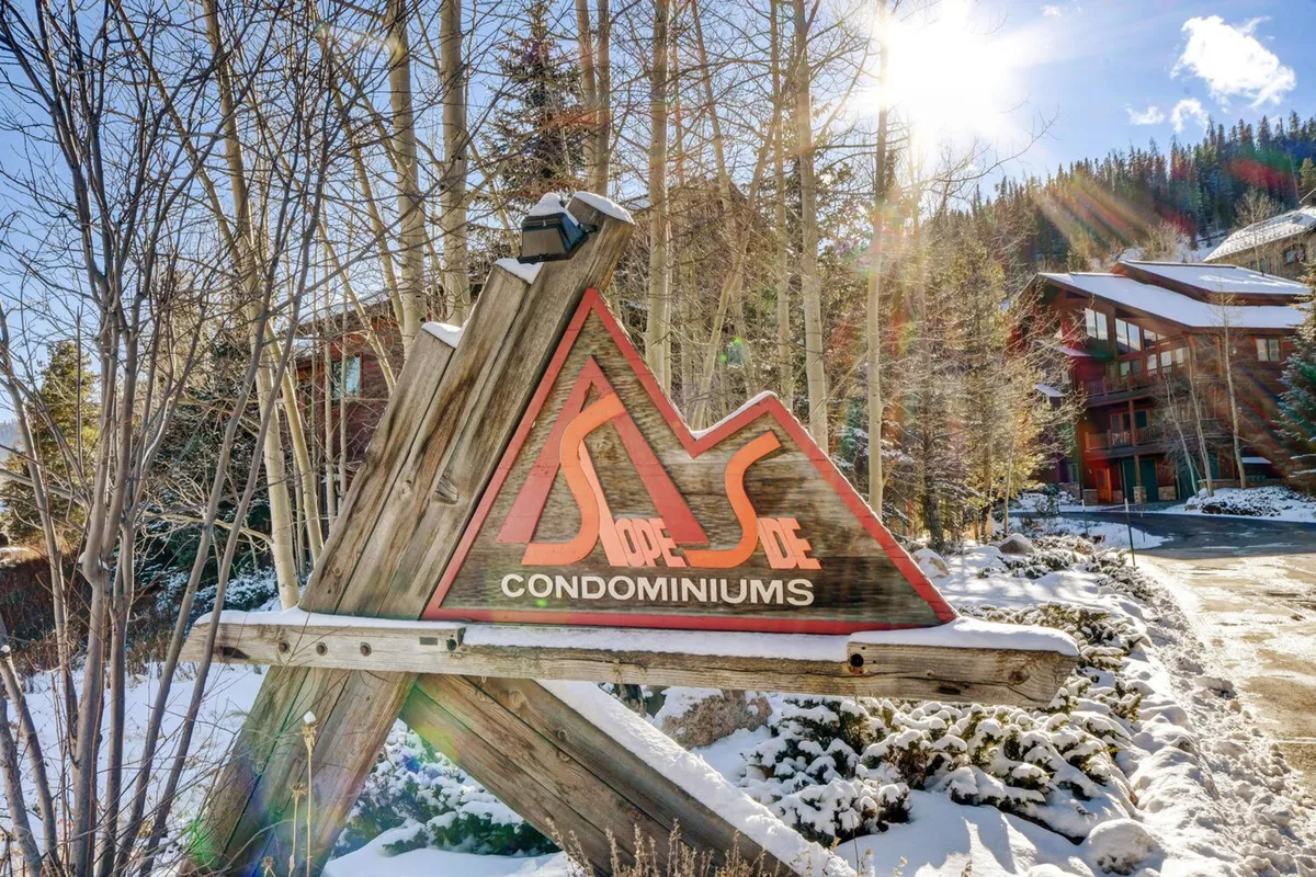 This exterior image shows the Slope Side Condominiums sign set against a snow-covered landscape, with surrounding trees and mountains in the background. The scenery emphasizes the property's proximity to outdoor adventures and the beauty of Colorado's winter environment.