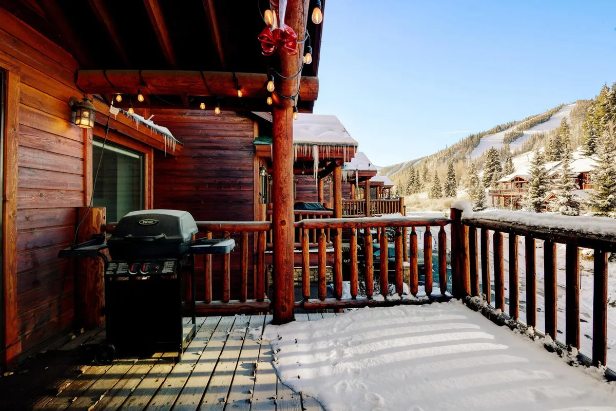 A spacious wooden deck features a gas grill positioned for easy outdoor cooking, with a wooden railing framing breathtaking views of the surrounding snow-capped mountains. The scene invites social gatherings and starlit grilling sessions.