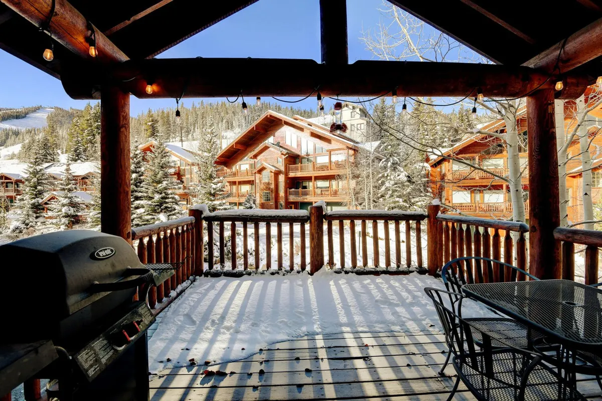 A cozy log cabin exterior featuring a snow-covered ground, a welcoming outdoor dining table with chairs, a grill, and a stunning backdrop of mountain views. The scene hints at stargazing opportunities and tranquil evenings spent in the fresh mountain air.