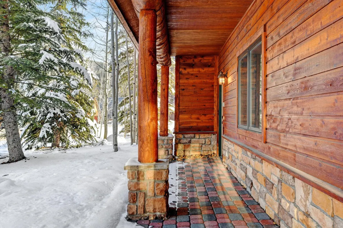 A snow-covered porch with a stone base and colorful pathway, framed by trees. The inviting wooden porch offers a place to relax and enjoy the wintry landscape, suggesting moments of quiet reflection or socializing with loved ones.