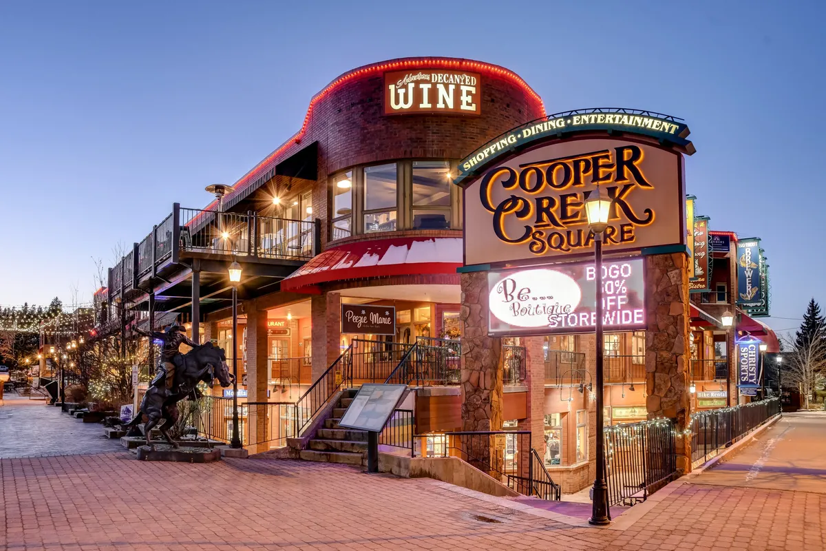 A lively streetscape featuring the Cooper Creek Square sign, outdoor seating, wine shop signage, and a prominent horse statue. People gather in the communal area, hinting at shopping, dining, and vibrant social happenings.