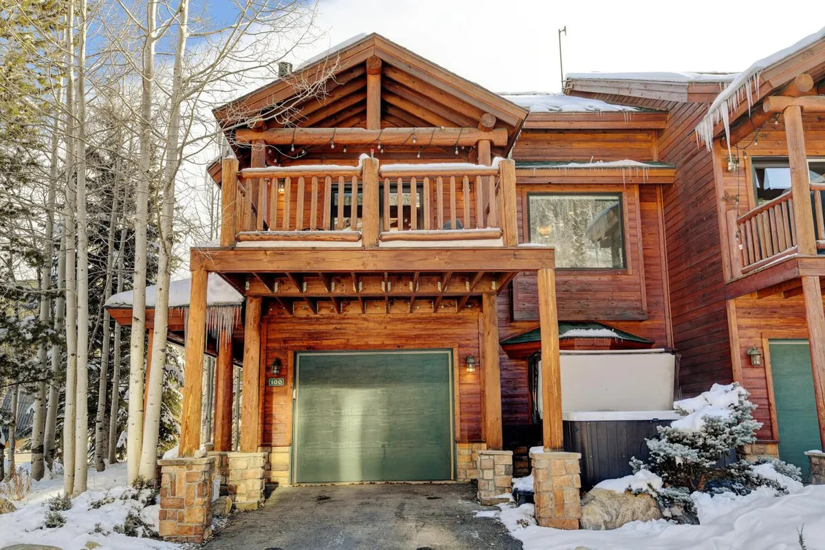 The property’s wooden balcony and green garage door are surrounded by a picturesque, snow-covered landscape. Stone accents and winter scenery evoke a serene, mountain hideaway feel, inviting guests to imagine cozy evenings and snowy adventures.
