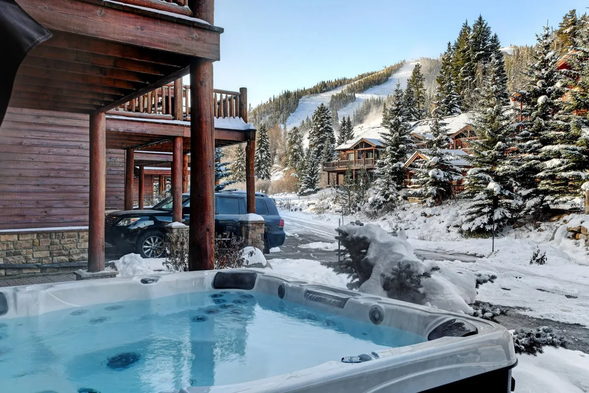 An inviting outdoor hot tub is nestled on a private patio, surrounded by snow-covered trees and wooden architecture. In the background, majestic mountains rise against the winter sky, creating a tranquil and festive alpine setting.
