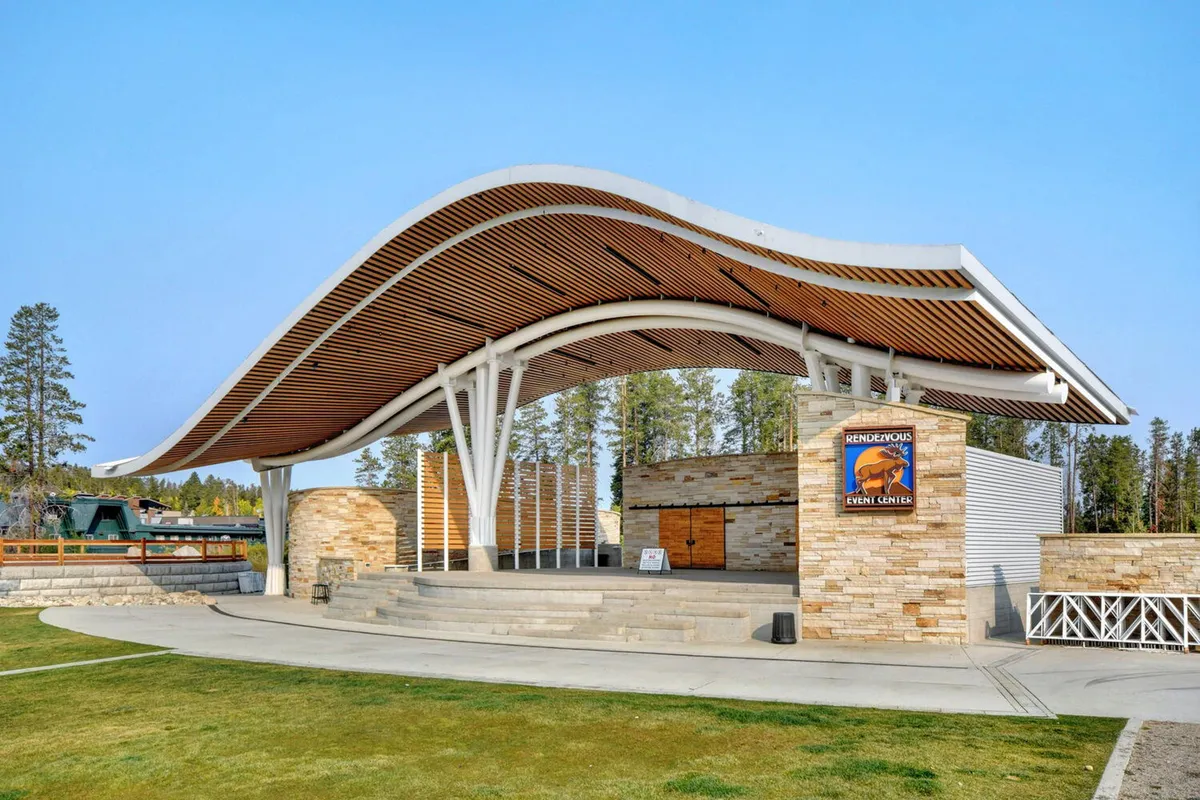 The exterior of an event center with a distinctive curved roof and stone facade, with signage indicating its role as a hub for community gatherings, outdoor concerts, and seasonal festivals. The building sits against a backdrop of clear skies and mountain scenery.