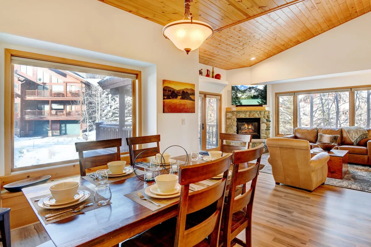This image presents an open-concept living and dining area with a spacious table set for a meal, comfortable seating, a fireplace, and large windows that frame scenic mountain views. The arrangement of furniture encourages gathering and socializing, while the fireplace and windows enhance the warm, inviting atmosphere.