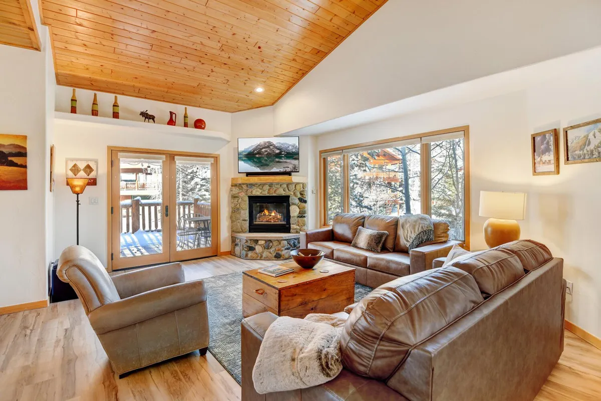 A cozy living area featuring a glowing fireplace, large windows with views, a wooden ceiling, comfortable sofas, and a coffee table. The room is ideal for relaxing after a day of mountain adventures, hosting game nights, or enjoying a warm beverage while taking in the scenery.